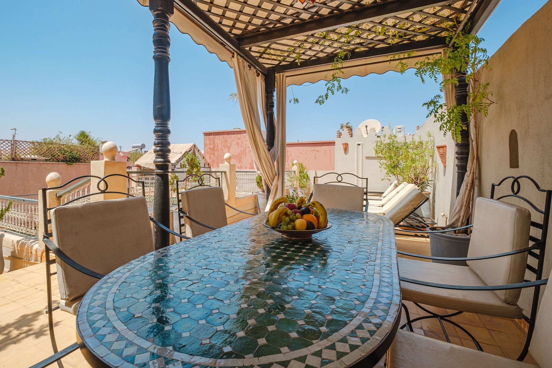 Rooftop dining area with mosaic table, cushioned chairs, pergola, and fruit bowl.