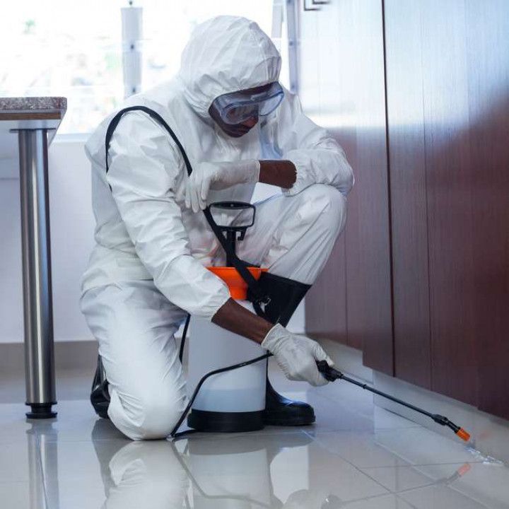 Worker in white protective suit using a sprayer to disinfect a floor indoors