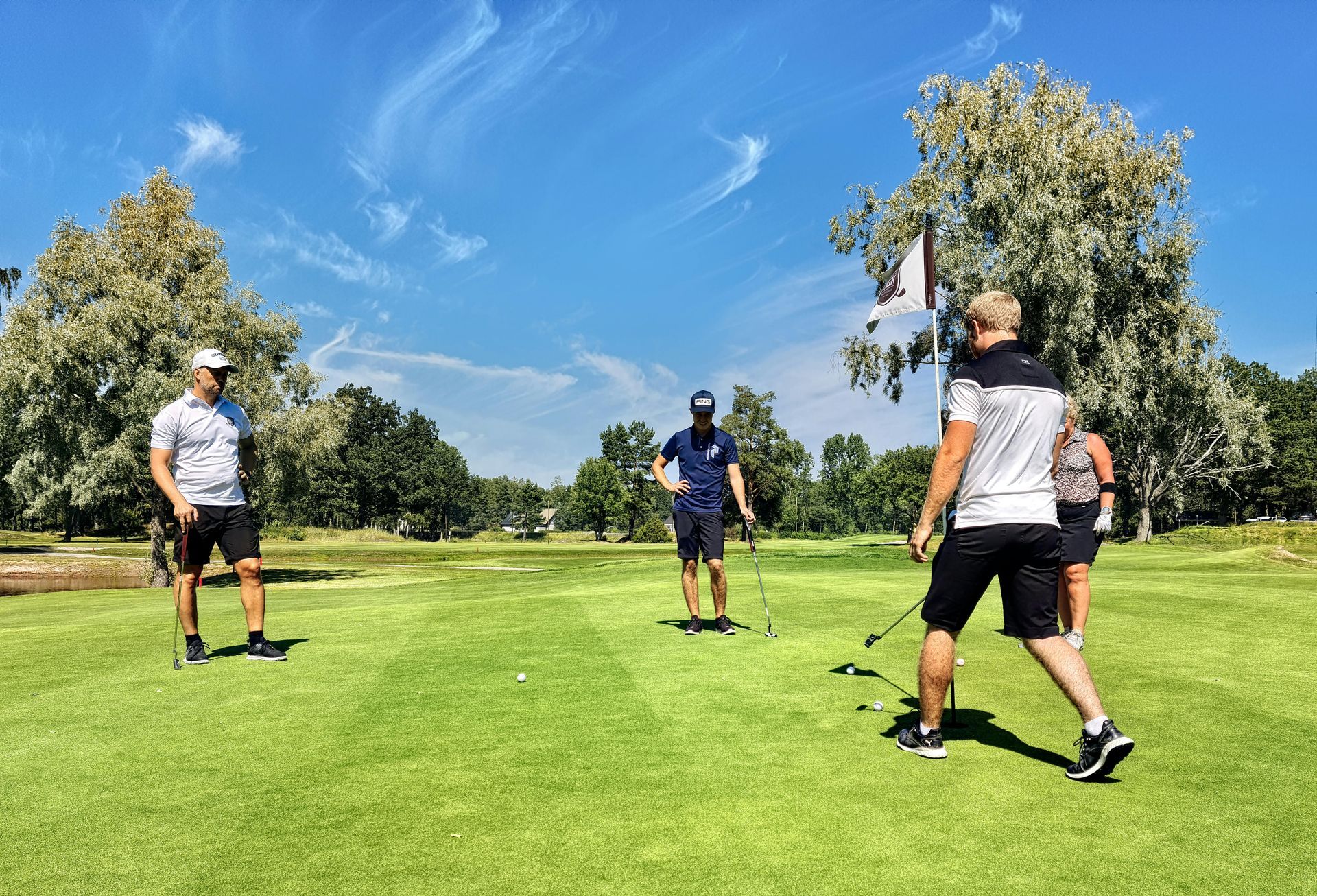 Four people in golf attire standing on a sunny putting green near a flag, preparing to play.