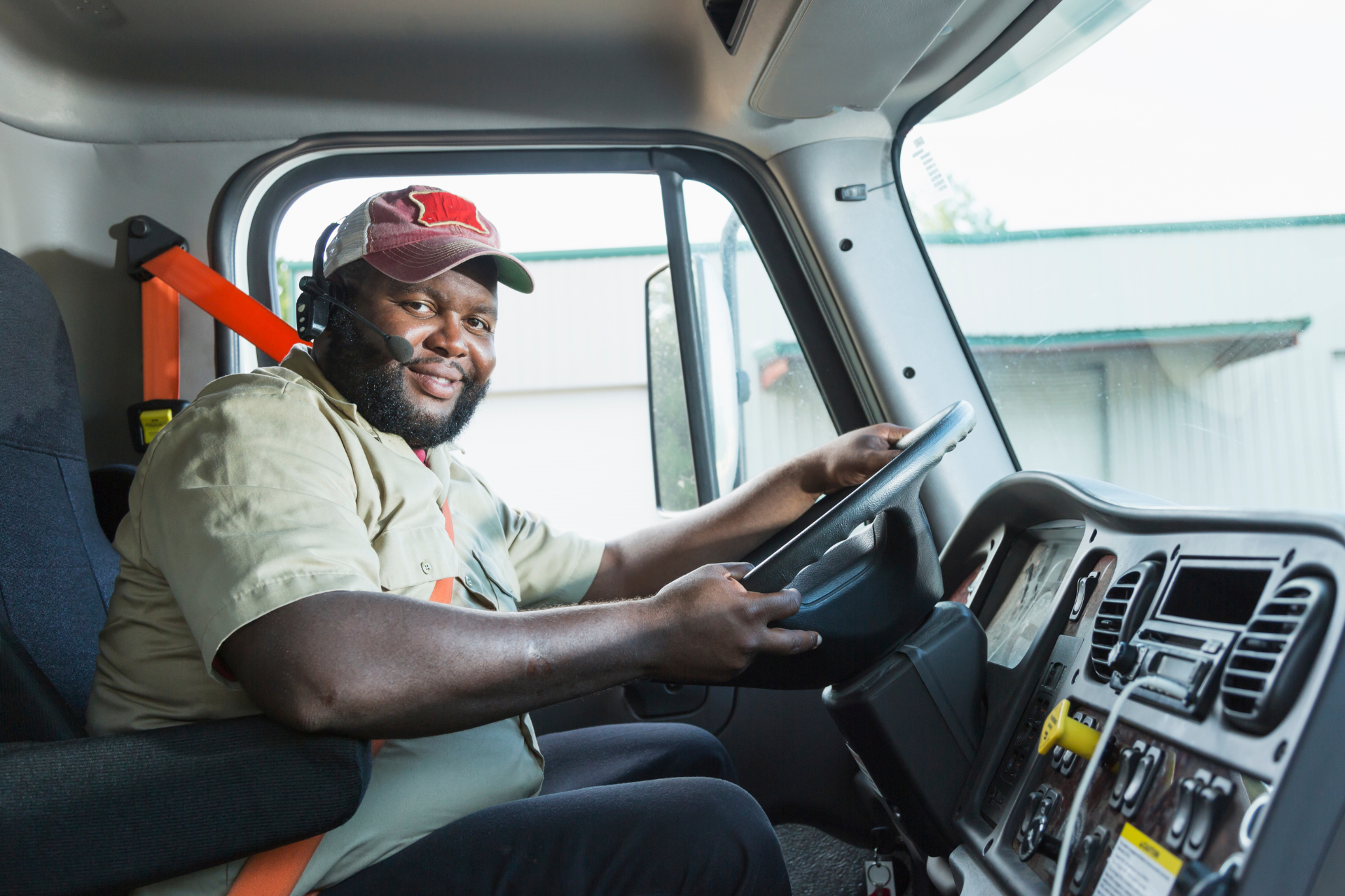Driver smiling in a truck cab, hands on the wheel, wearing a red cap and beige shirt.