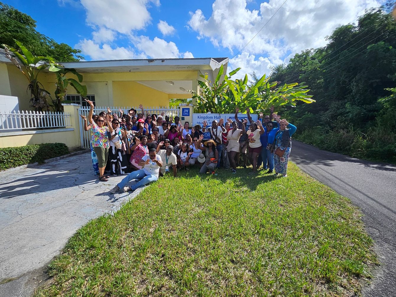 Group of people waving in front of a yellow building with green vegetation, under a blue sky.