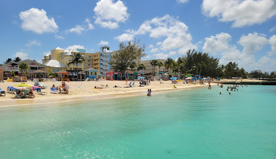 Beach scene with turquoise water, white sand, people swimming and sunbathing; buildings and blue sky with clouds in the background.