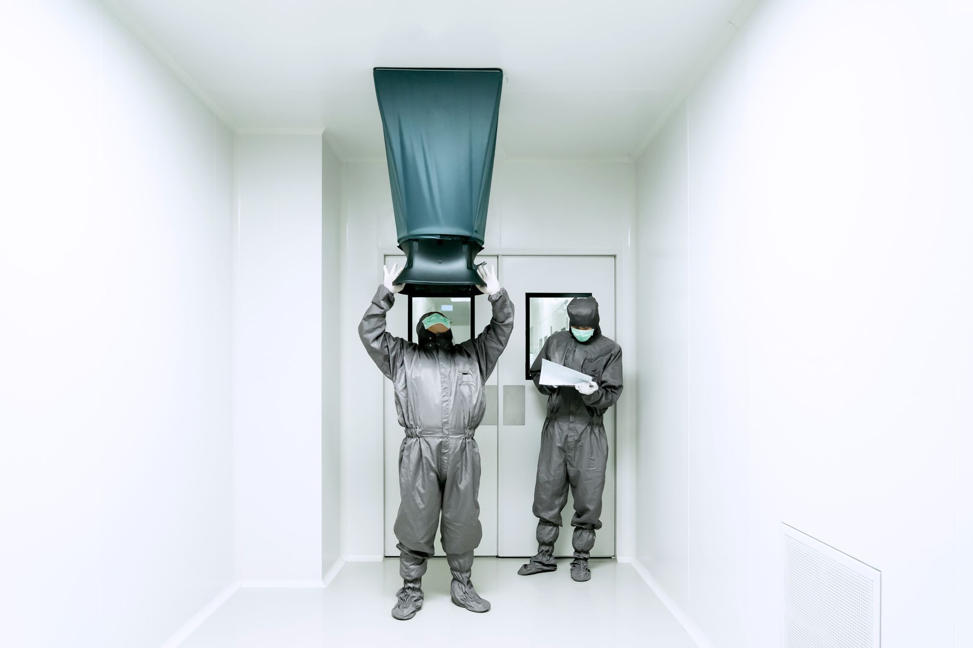 Two people in protective suits in a white room; one holds a large black air vent. A door is behind them.