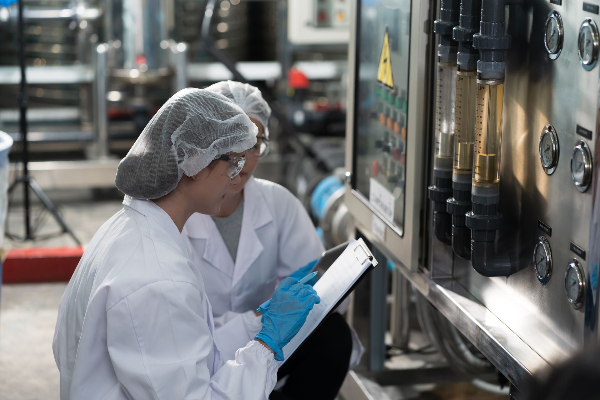 Two people in lab coats inspect equipment in a factory setting, one holding a clipboard.