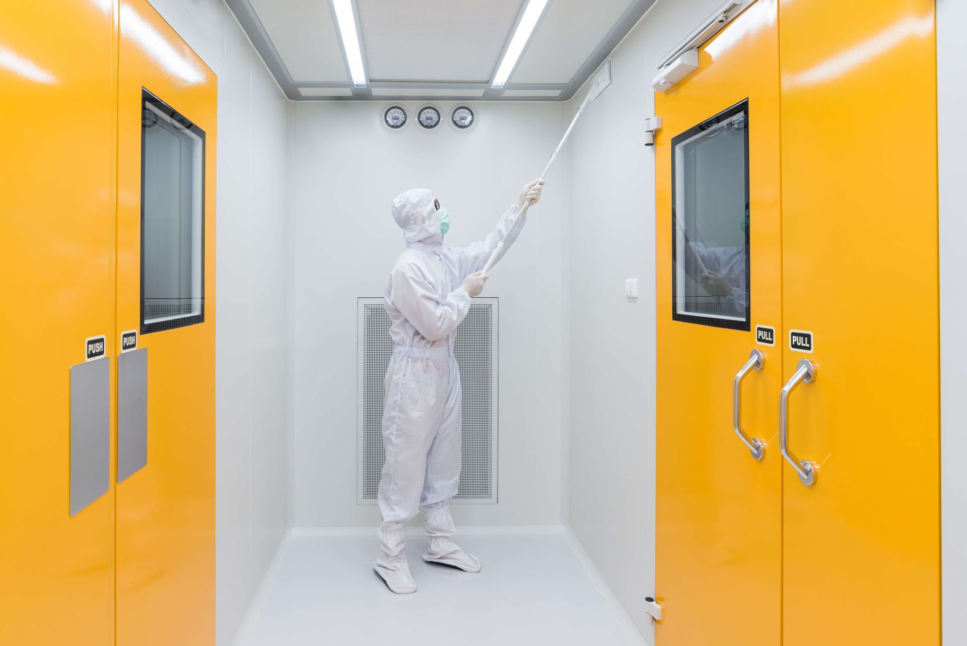 Person in cleanroom suit cleaning ceiling. Bright yellow doors and white walls.