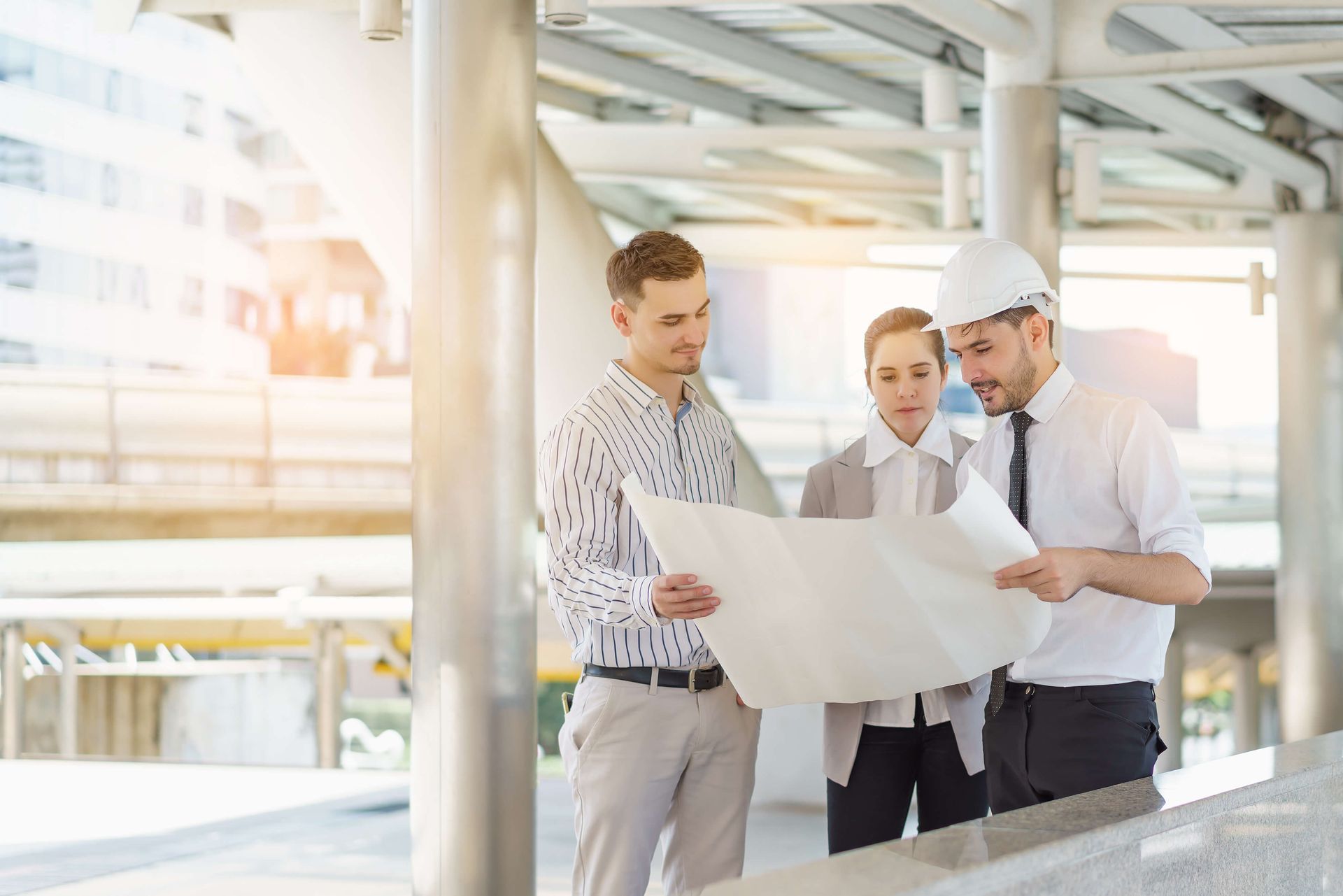 Three people looking at a blueprint outdoors near a modern building. One wears a hardhat.