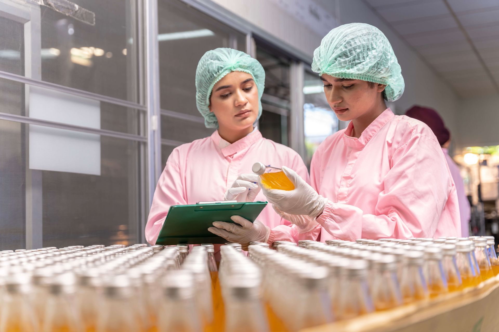 Two workers in pink lab coats and hairnets inspecting bottles of liquid on a production line.