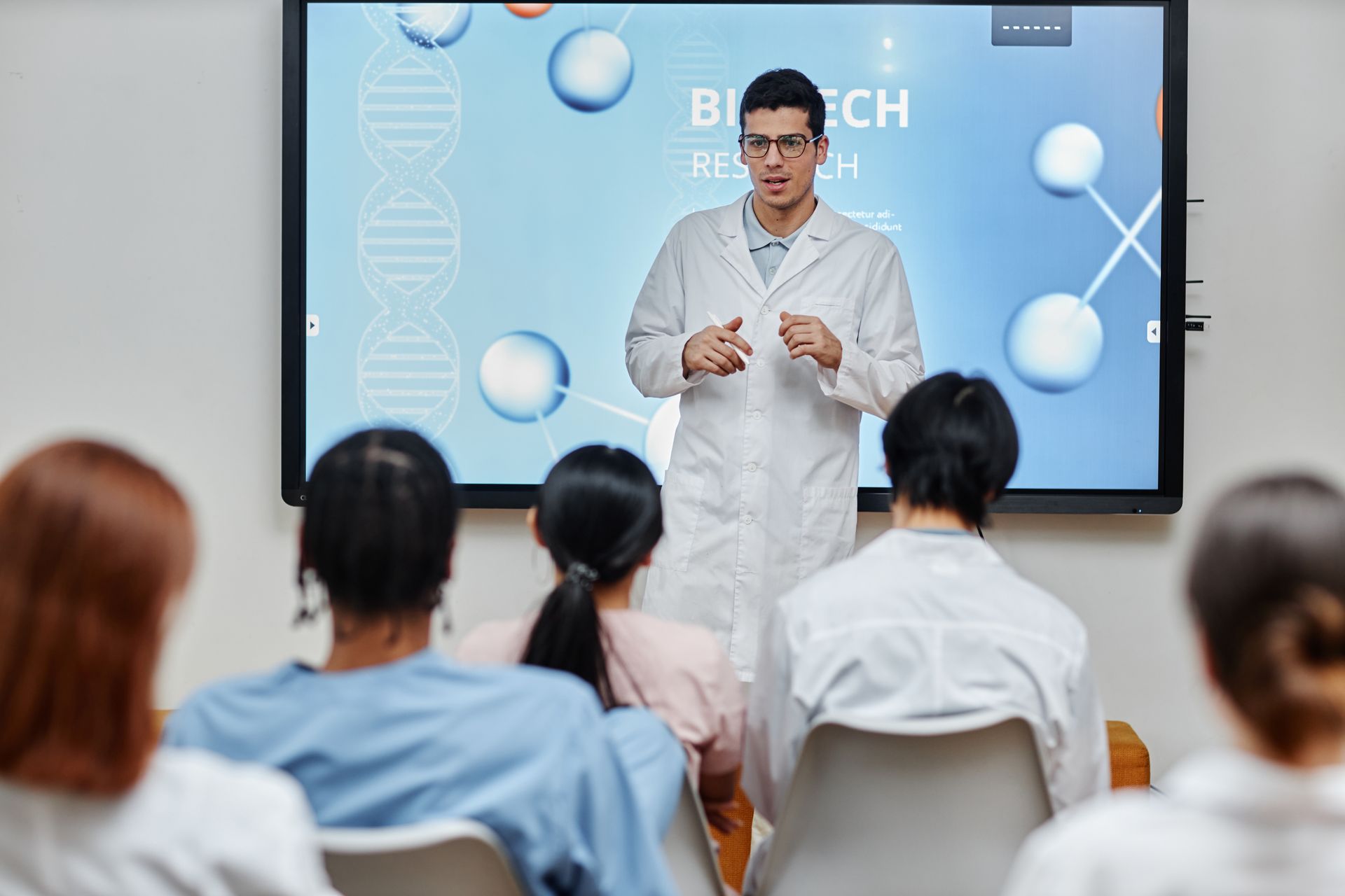 Scientist in lab coat presents to audience in front of Biotech research screen.