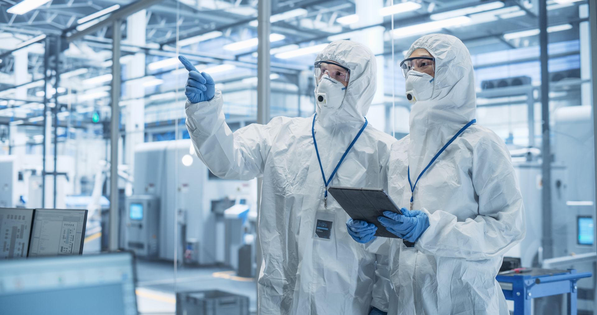 Two workers in white protective suits inside a clean room, one pointing, the other holding a tablet.