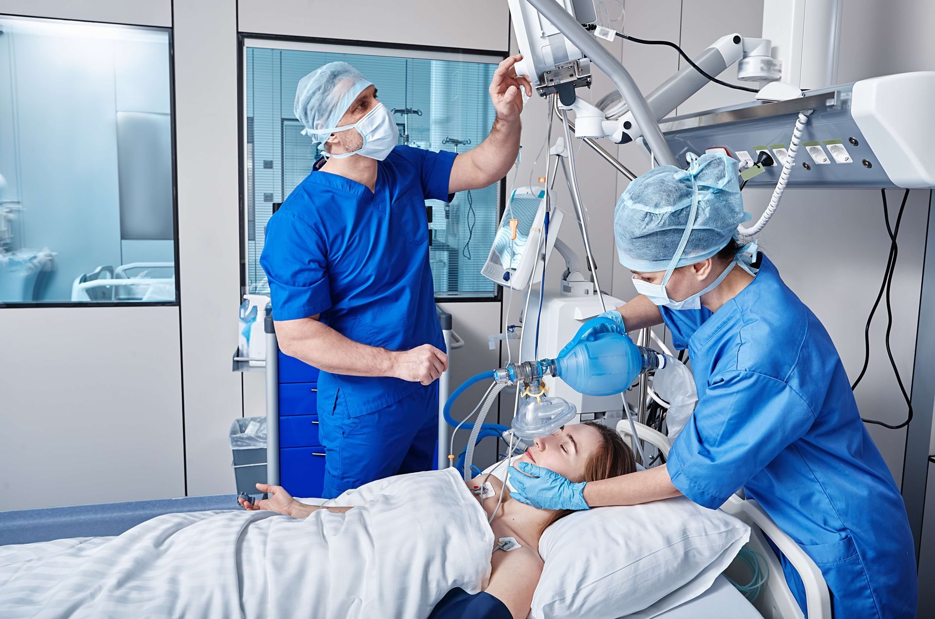 Two medical professionals assist a patient on a hospital bed. One adjusts equipment, the other holds a breathing mask.