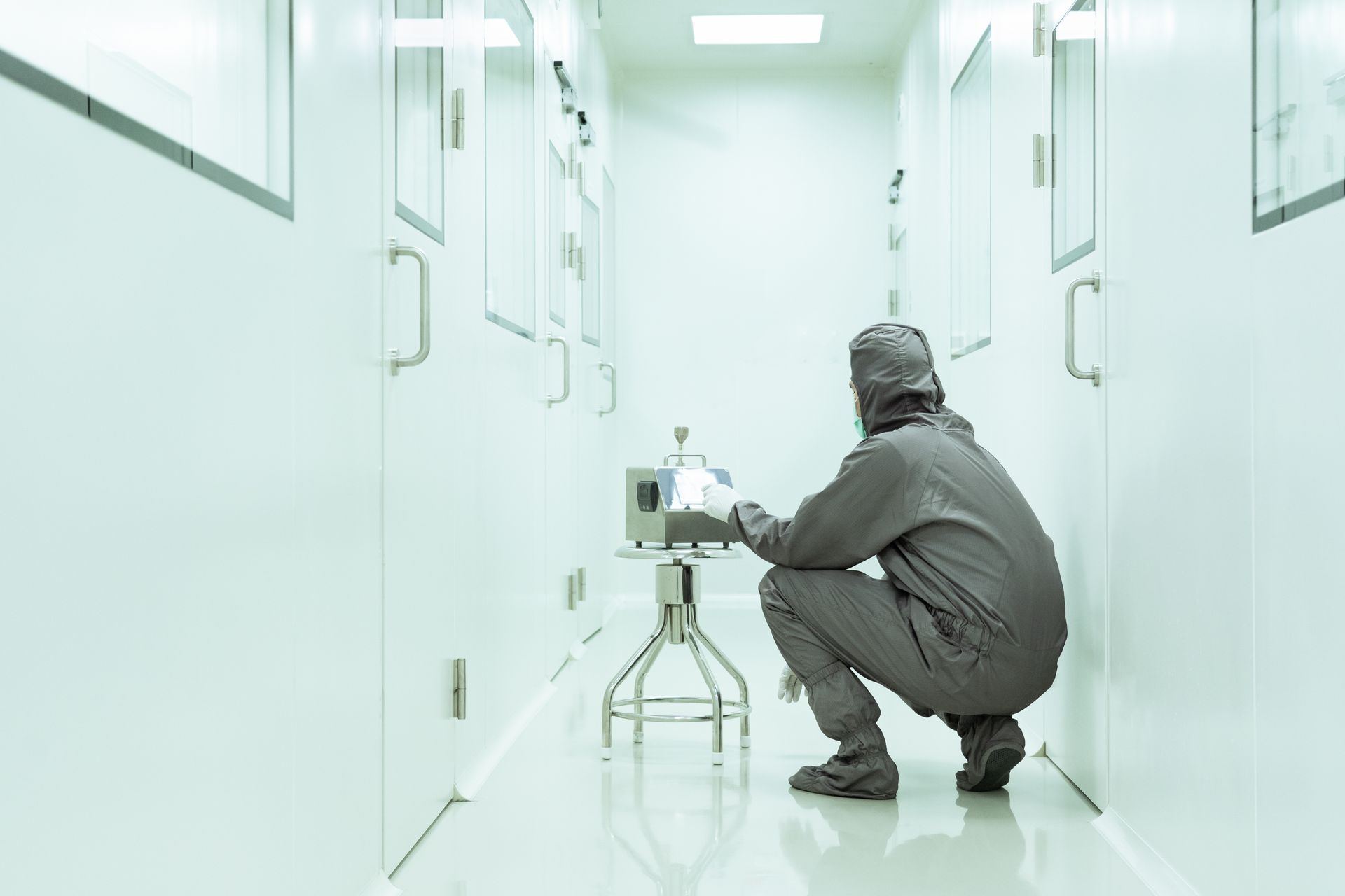 Person in sterile suit crouches in lab hallway, operating equipment near a door.