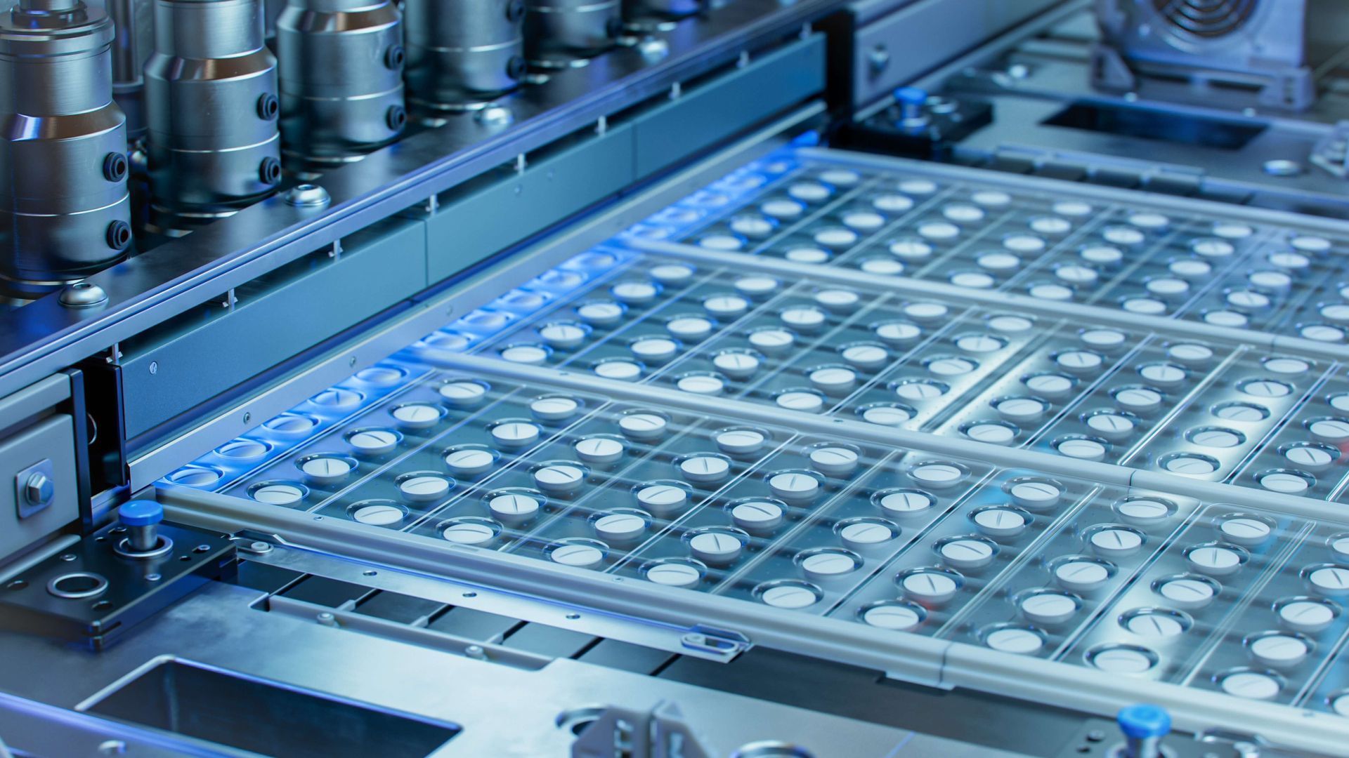 Automated manufacturing equipment with rows of small, circular white components arranged on a grid in a sterile blue light.