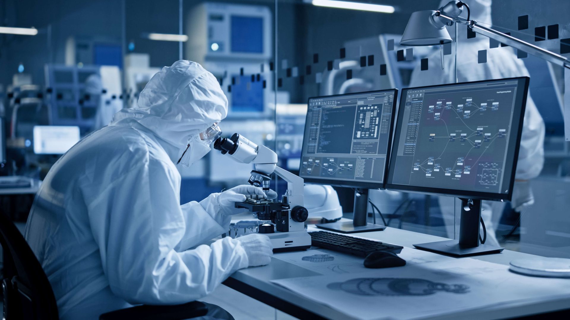 Person in a protective suit using a microscope in a laboratory, looking at two computer screens.