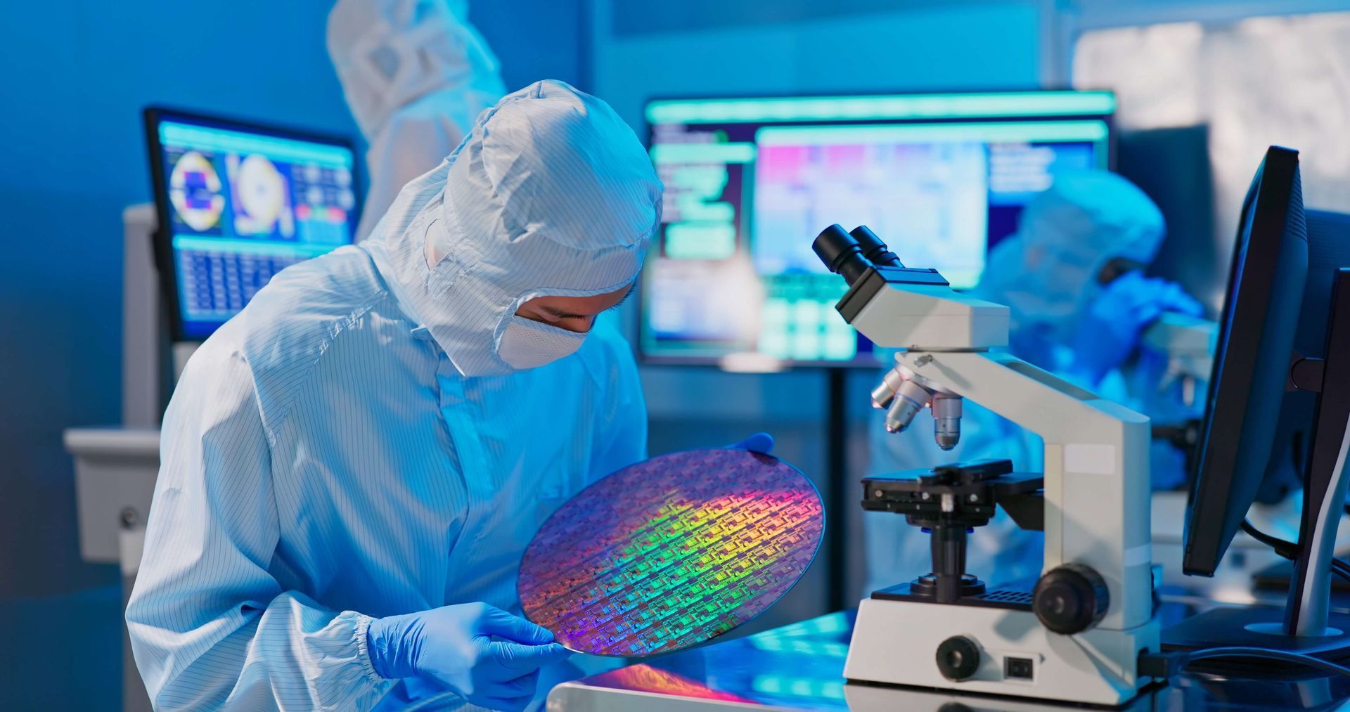 Scientist in cleanroom suit examines a silicon wafer under a microscope in a laboratory, another person in the background.