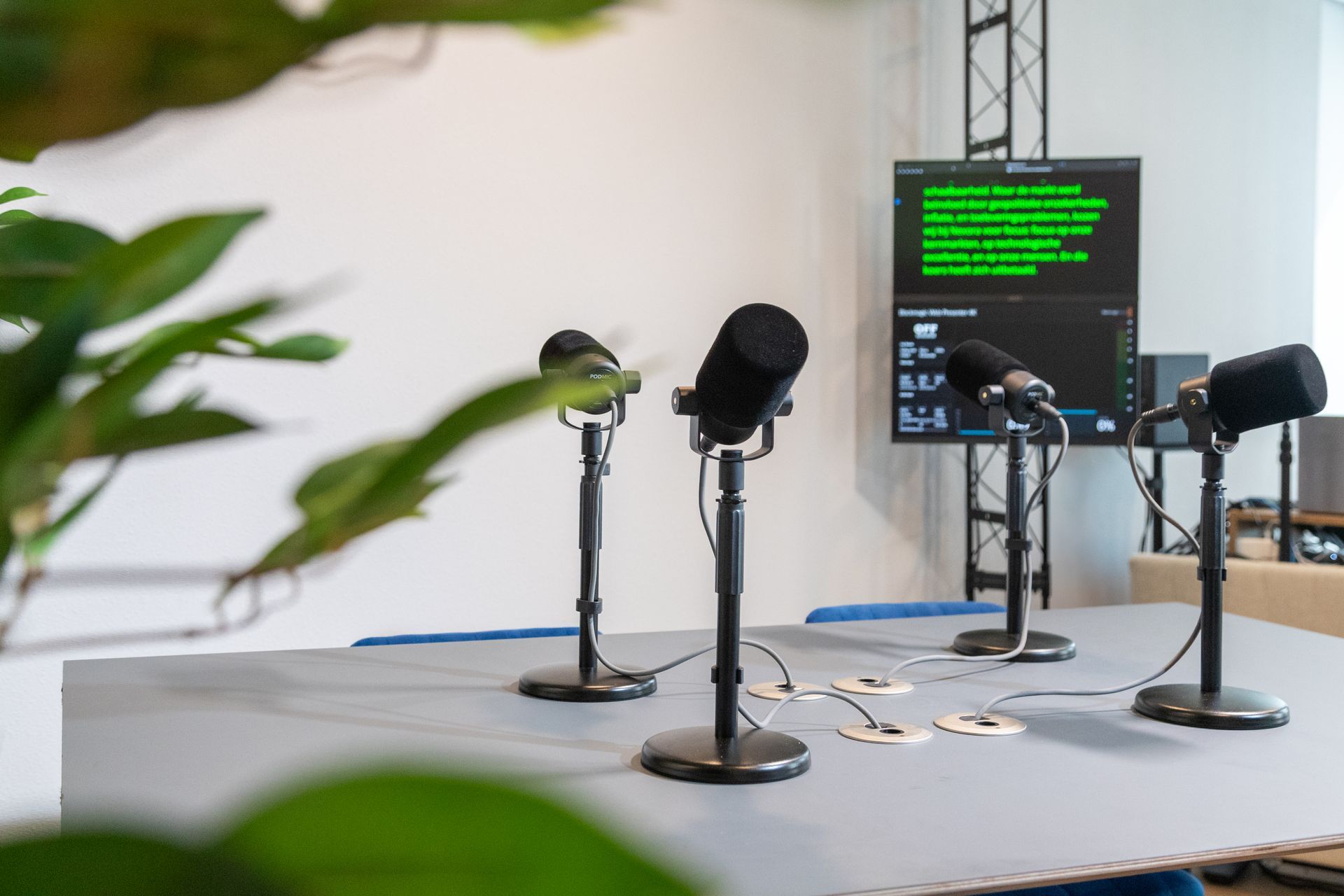Vier microfoons staan op een tafel opgesteld voor opnames, met daarachter een scherm waarop tekst wordt weergegeven.