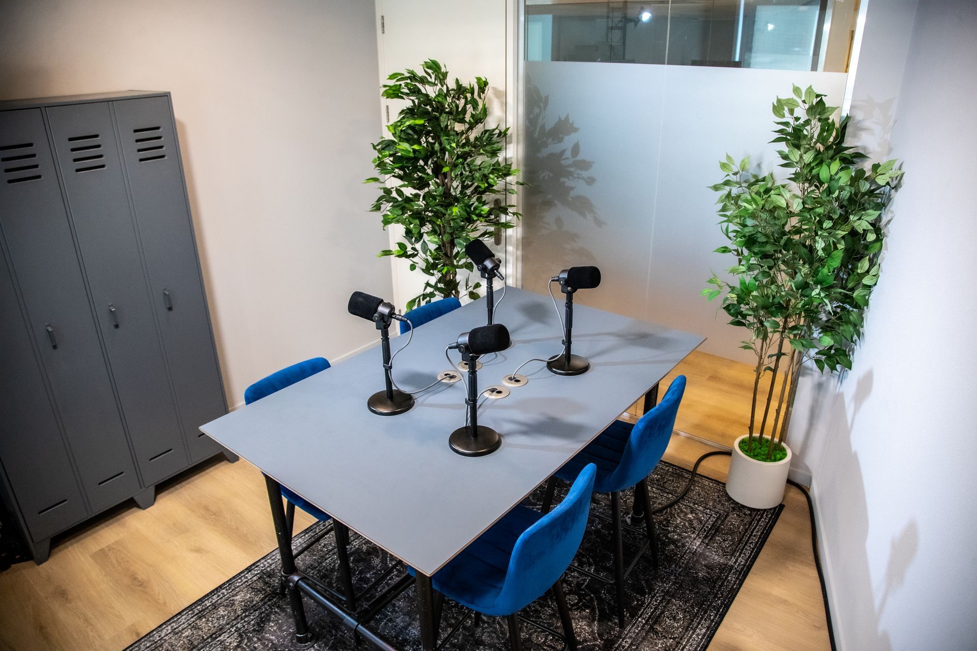 Podcaststudio met microfoons op tafel, blauwe stoelen, planten en een grijs kluisje.