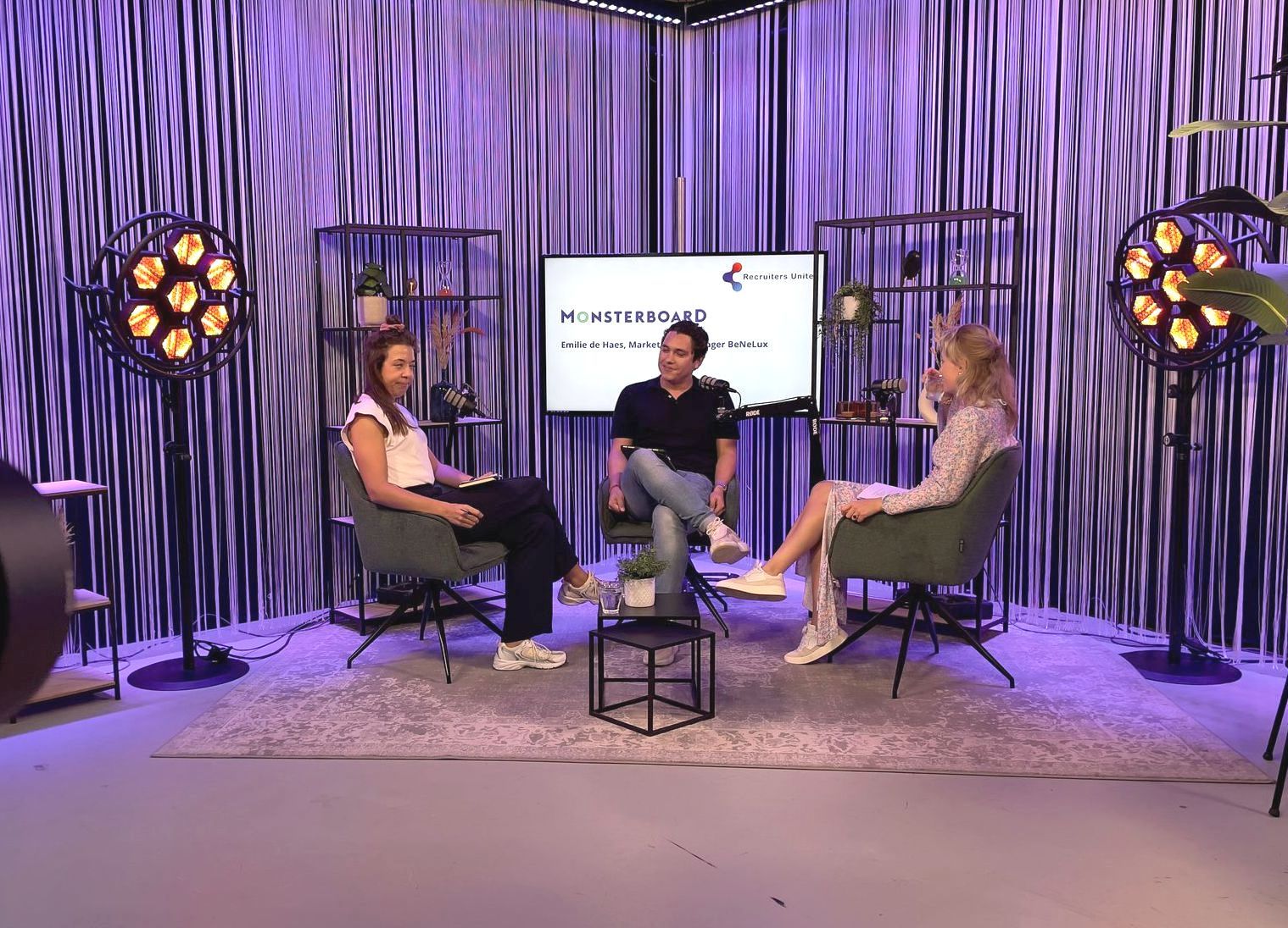 Drie mensen zitten in een tv-studio op stoelen te praten. Tegen een paars verlichte achtergrond.