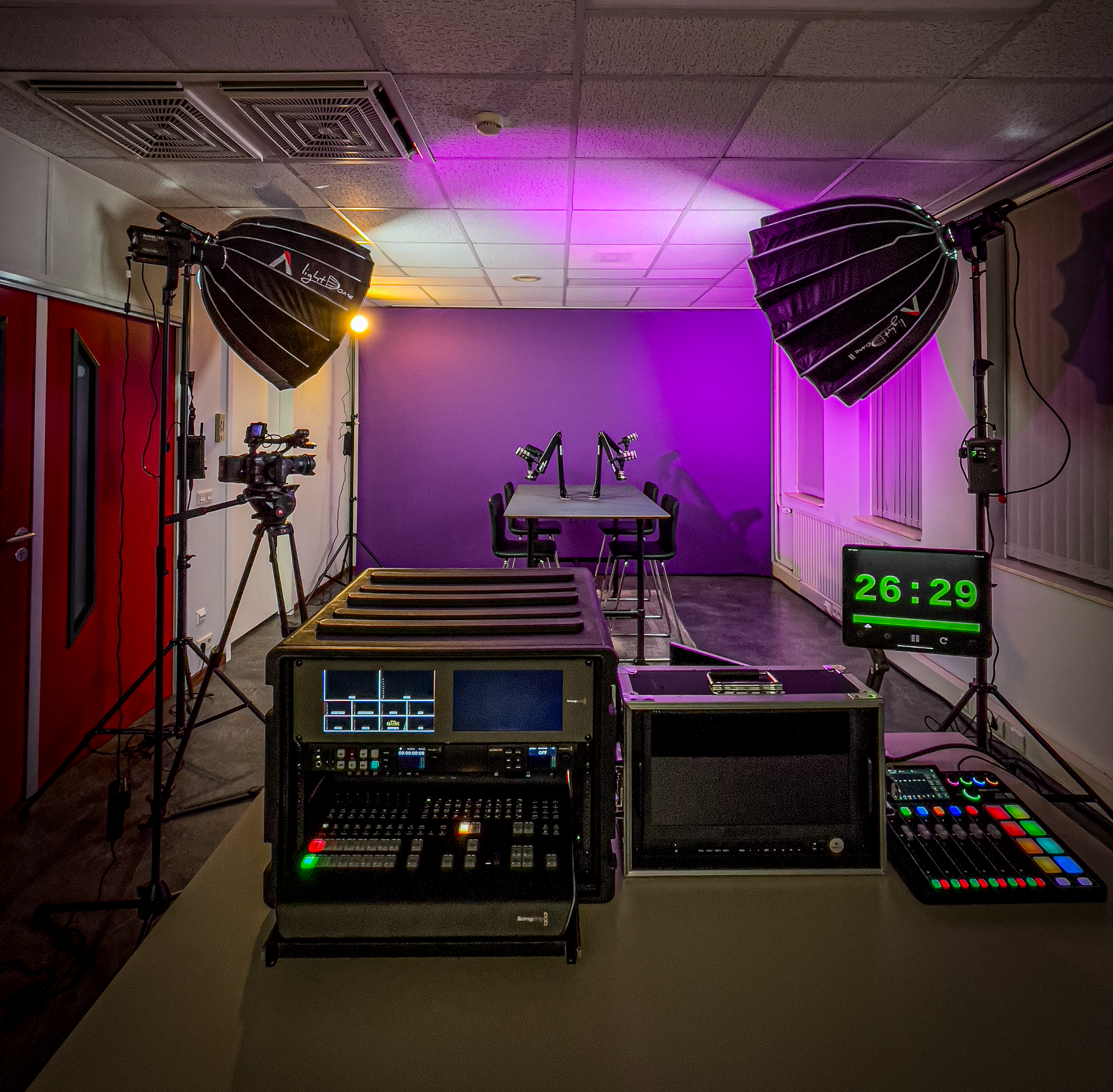 Een studio met opnameapparatuur, paarse en witte verlichting en een tafel met twee microfoons.