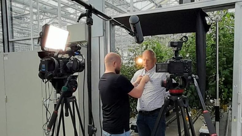 Twee mannen filmen een video in een kas. De een trekt het shirt van de ander recht, vlakbij camera's en verlichting.