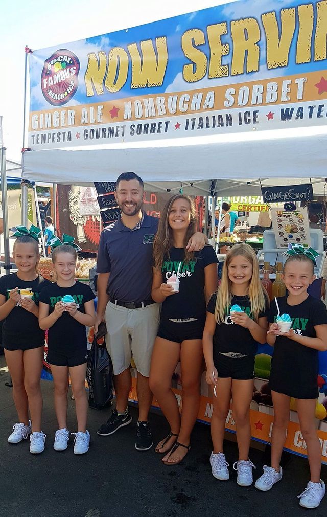 A man and five children in matching shirts stand smiling and holding treats in front of a Tropical Sno concession stand.