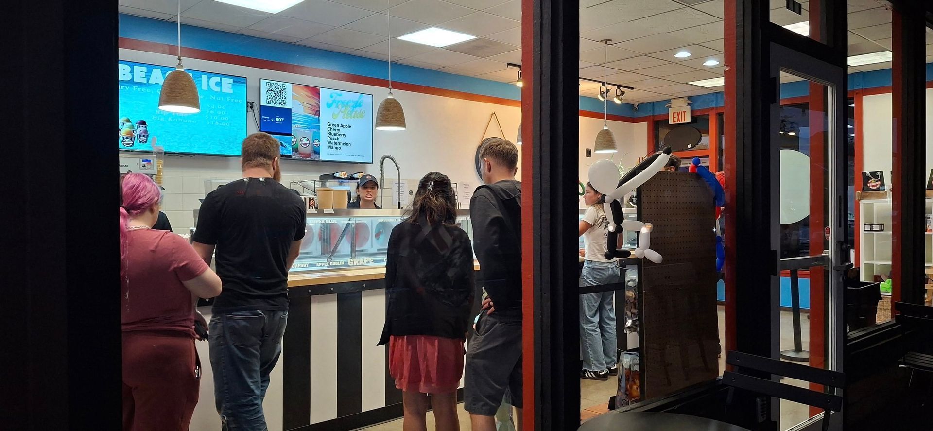 A group of people standing in a line waiting to be served at an ice cream shop counter with menu boards visible.