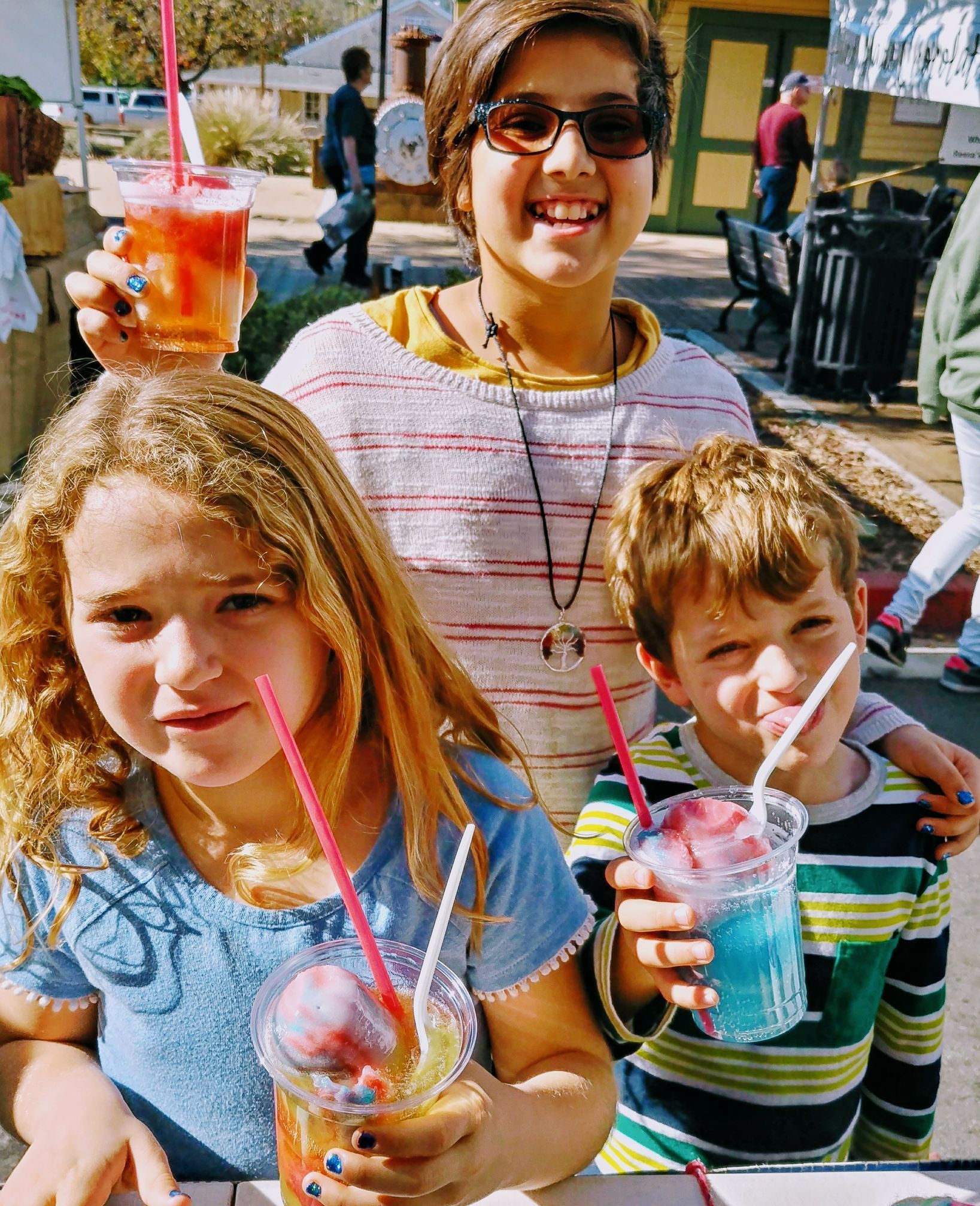 Three children holding colorful drinks with straws, smiling outdoors in a sunny setting.