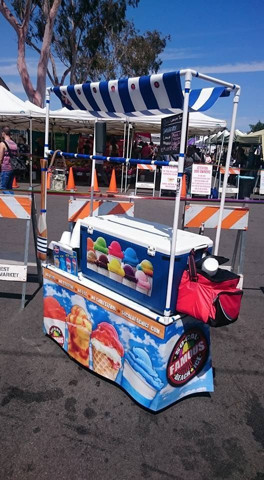 A portable ice cream cart with a blue and white striped canopy, parked outdoors in a public area.