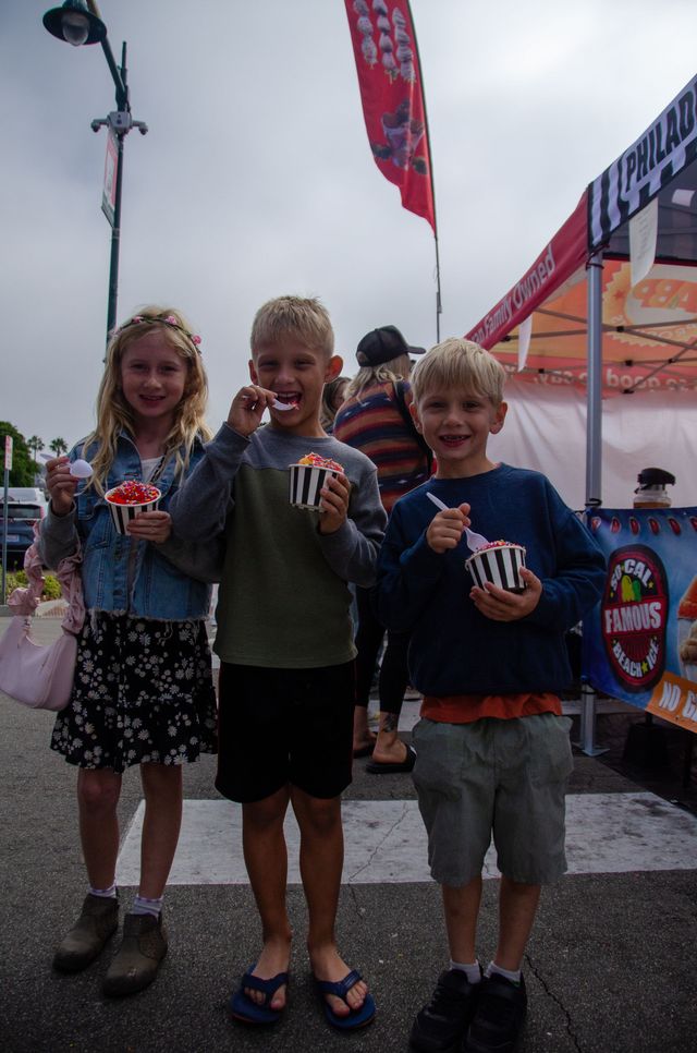 Three children stand outdoors in front of a food stall, smiling and holding cups of shaved ice with plastic spoons.