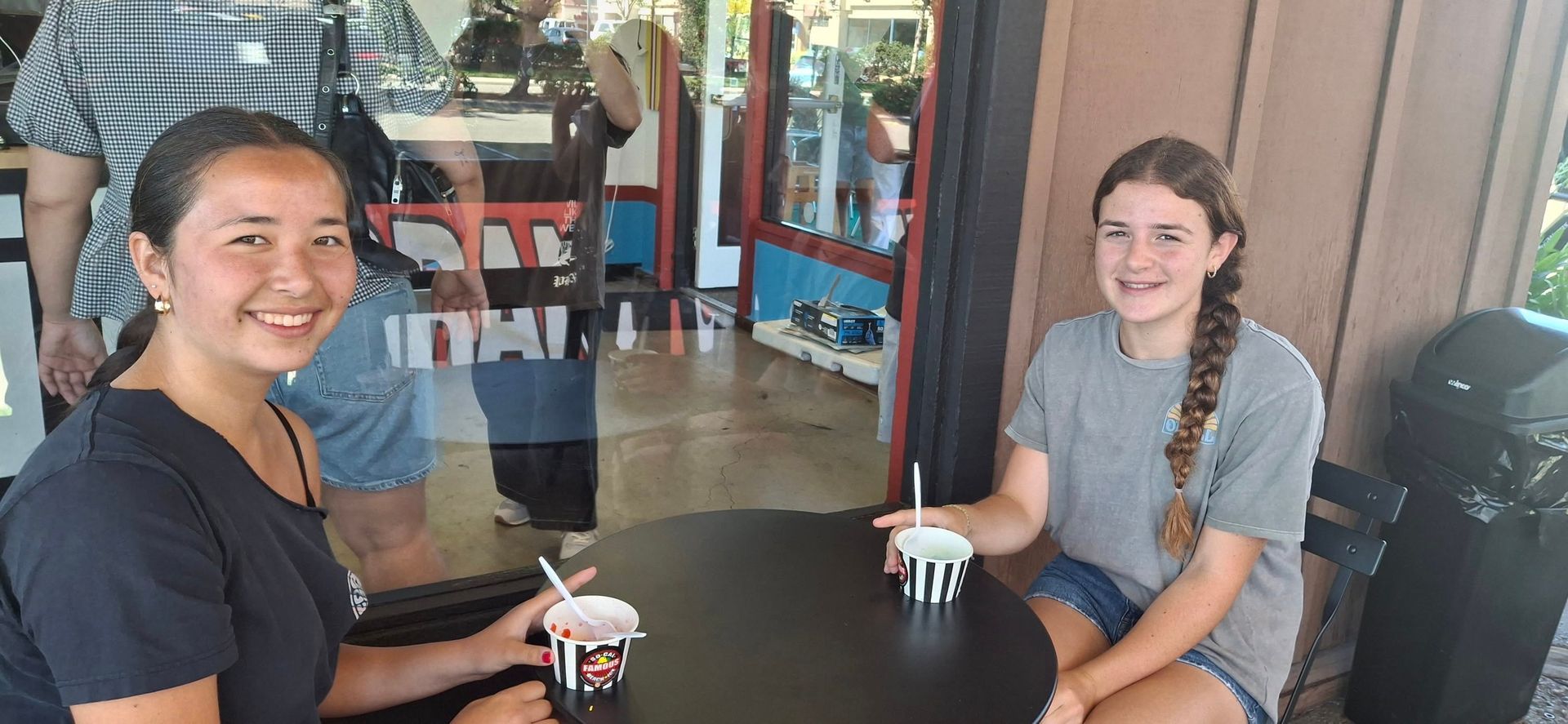 Two individuals smile while sitting at an outdoor table with small cups of frozen treats near a storefront.