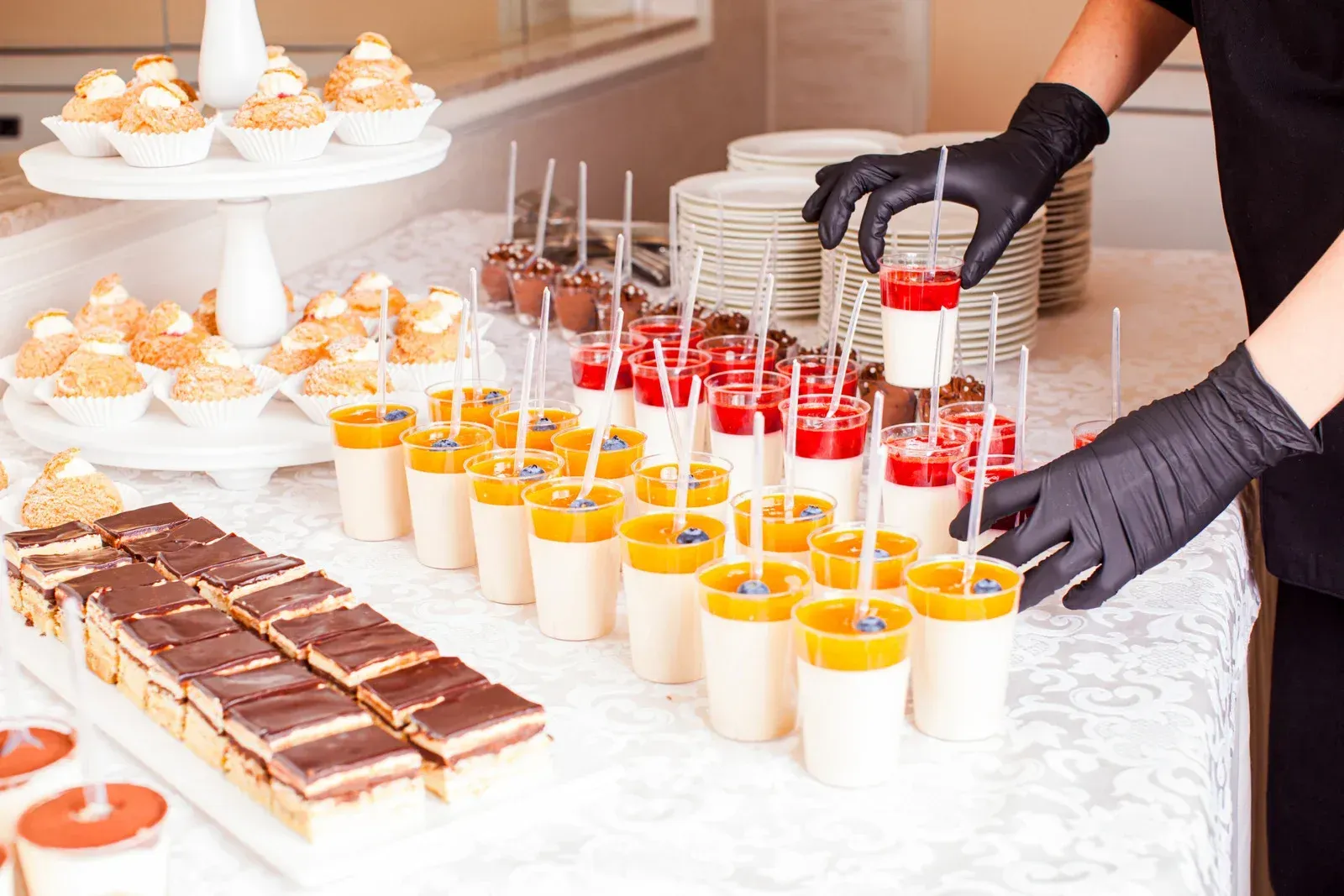 A person wearing black gloves arranges small dessert cups and pastries on a white tablecloth.