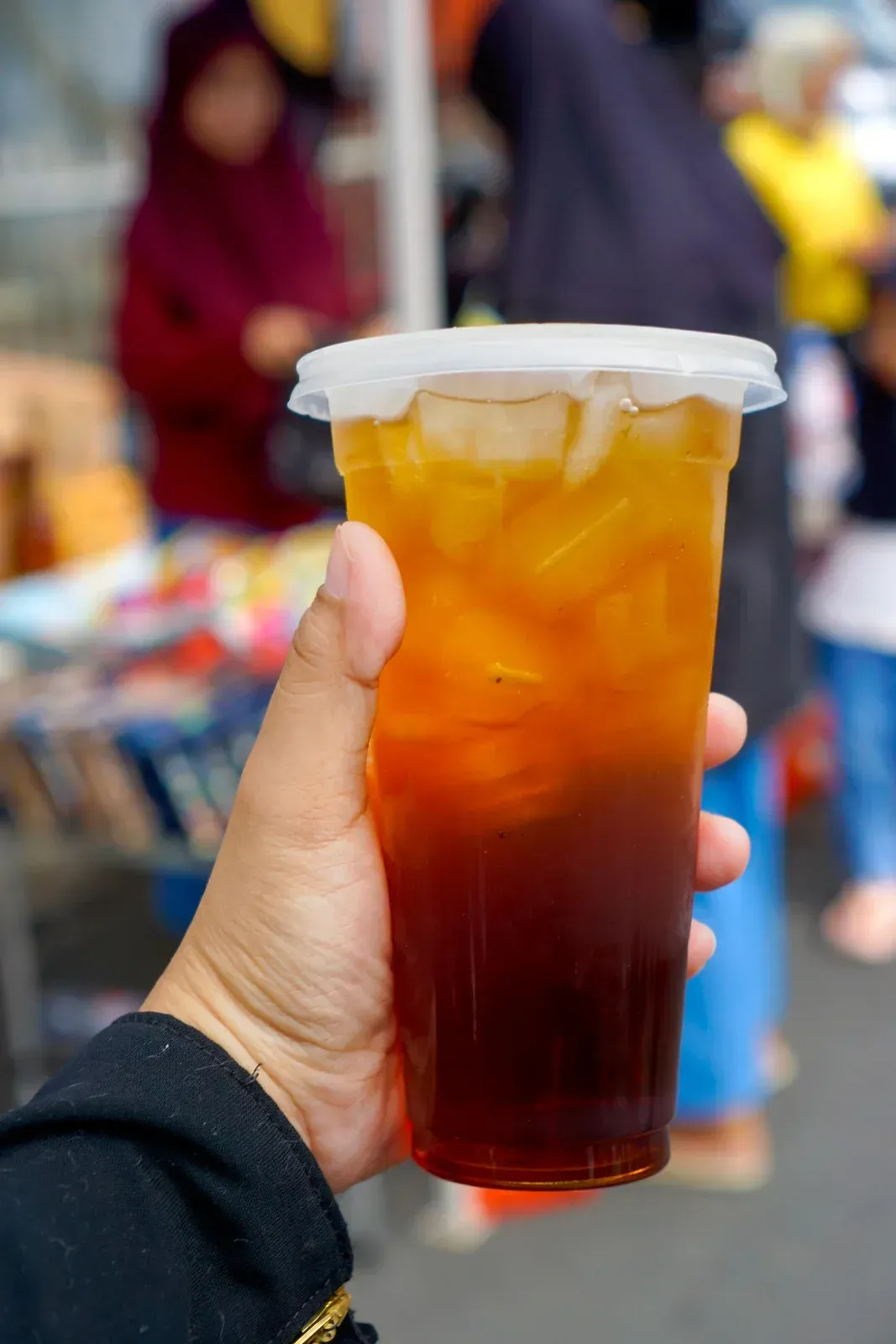 A hand holds a plastic cup of iced tea with a gradient color from dark amber at the bottom to light gold at the top.