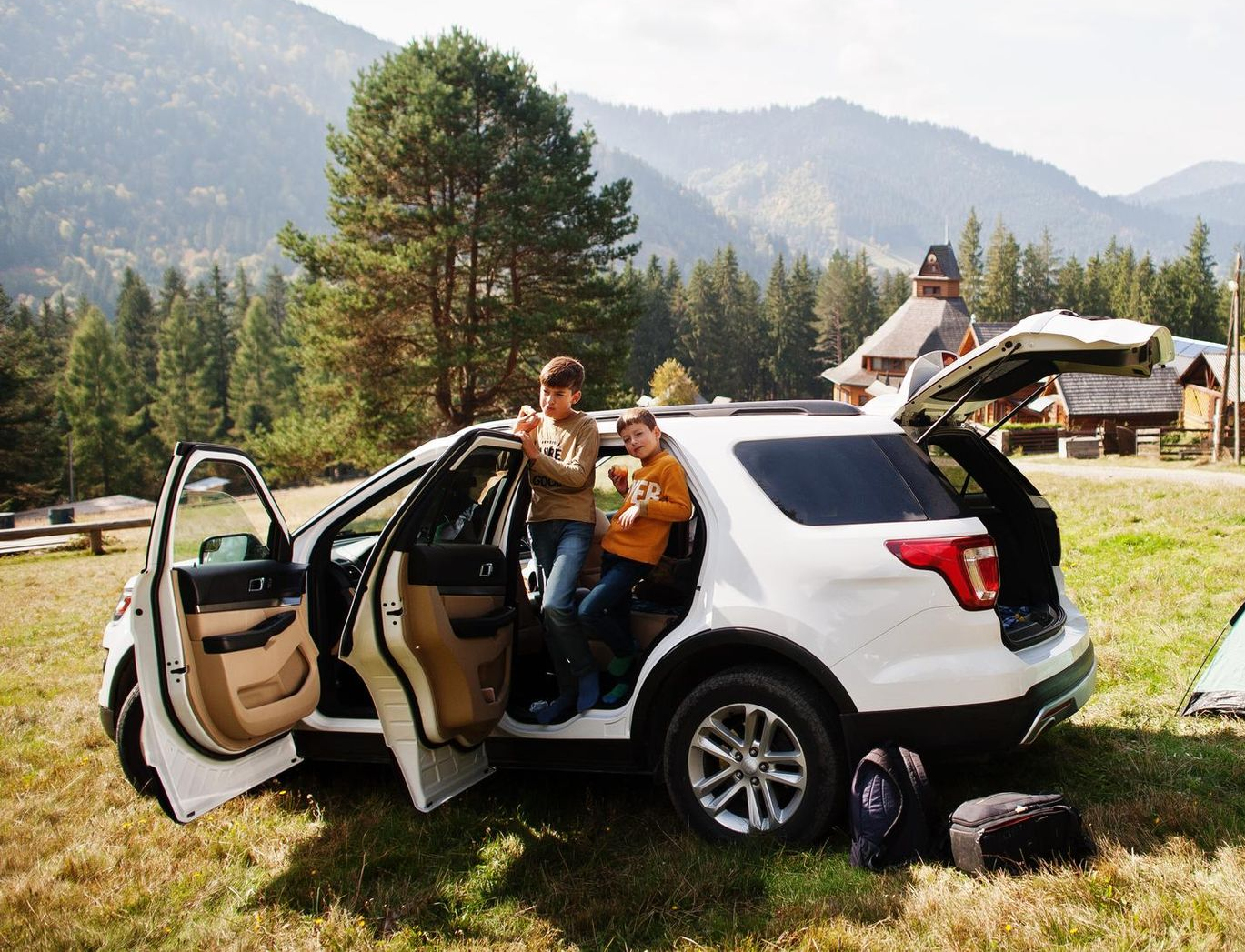 Two people sit in an open white SUV in a grassy mountain campsite, with an open door and trees behind them.