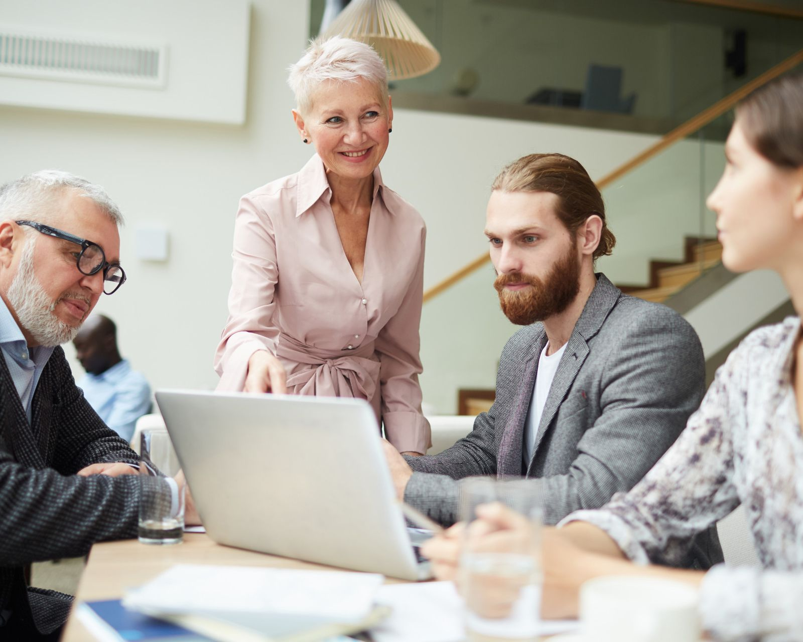 Business team in a meeting around a laptop, with one person standing and others seated in a bright office.