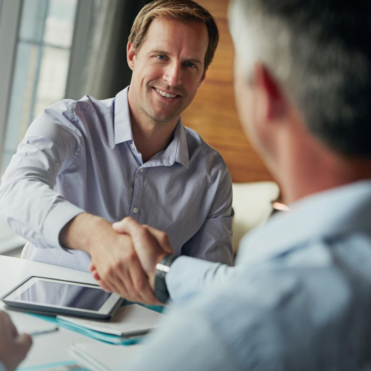 Two men shaking hands across a table in a business meeting, with a tablet nearby.