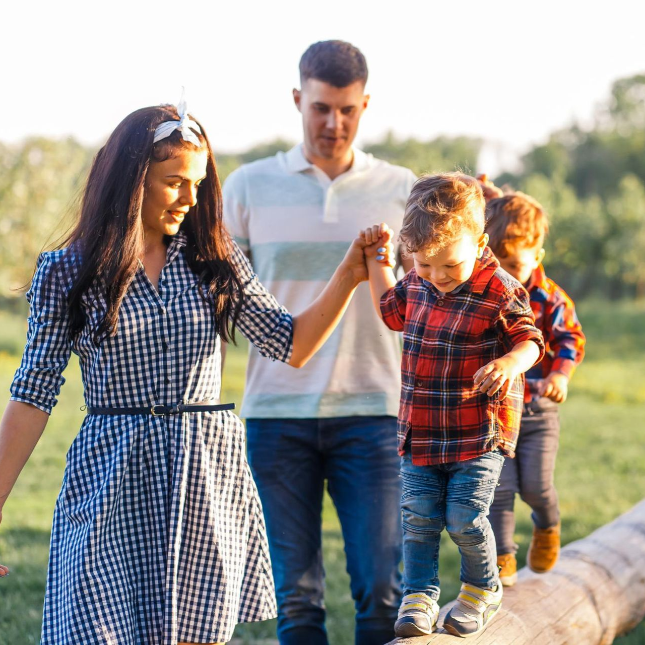 Family walking along a log on a sunny path, holding hands in a grassy field.