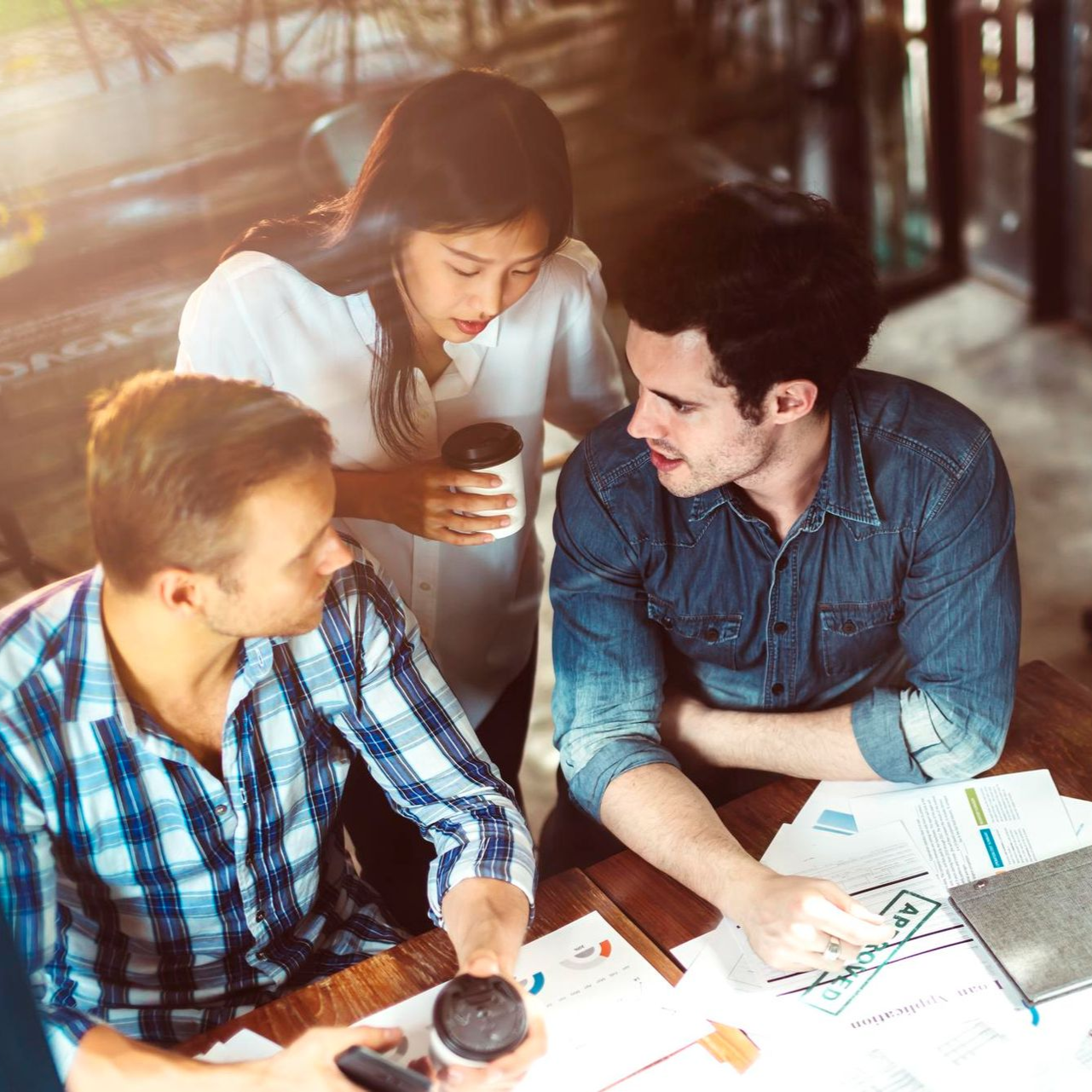 Three coworkers in a bright office review documents around a table with coffee cups and a laptop.