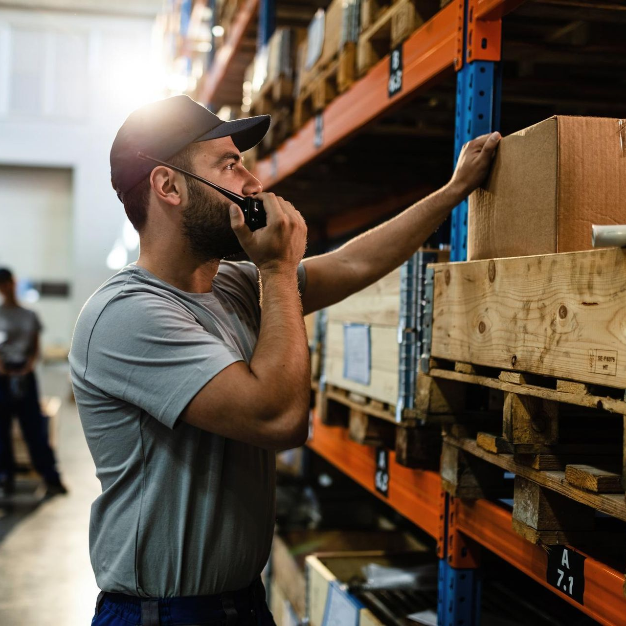 Warehouse worker speaking on a walkie-talkie while reaching for a box on a storage shelf.