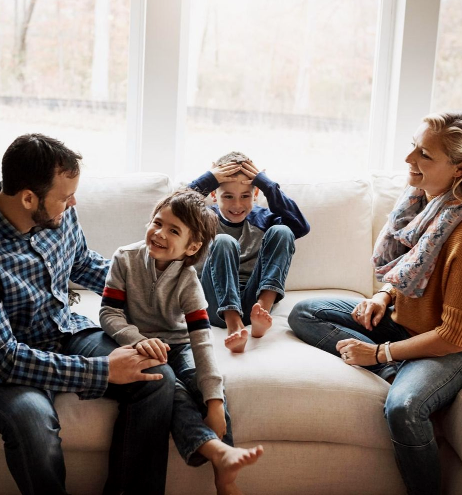 Family sitting together on a couch in a bright living room, smiling and talking.