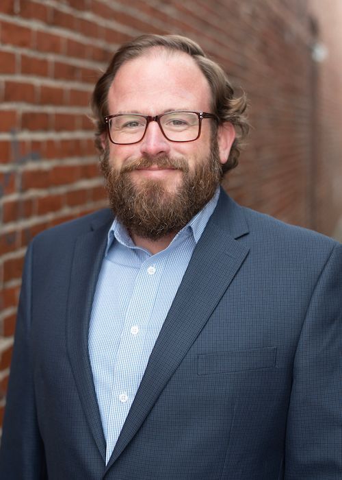 Man in glasses and blue blazer standing against a brick wall, smiling slightly