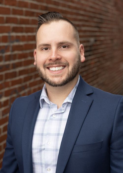 Smiling person in a navy blazer and light blue shirt standing against a brick wall.