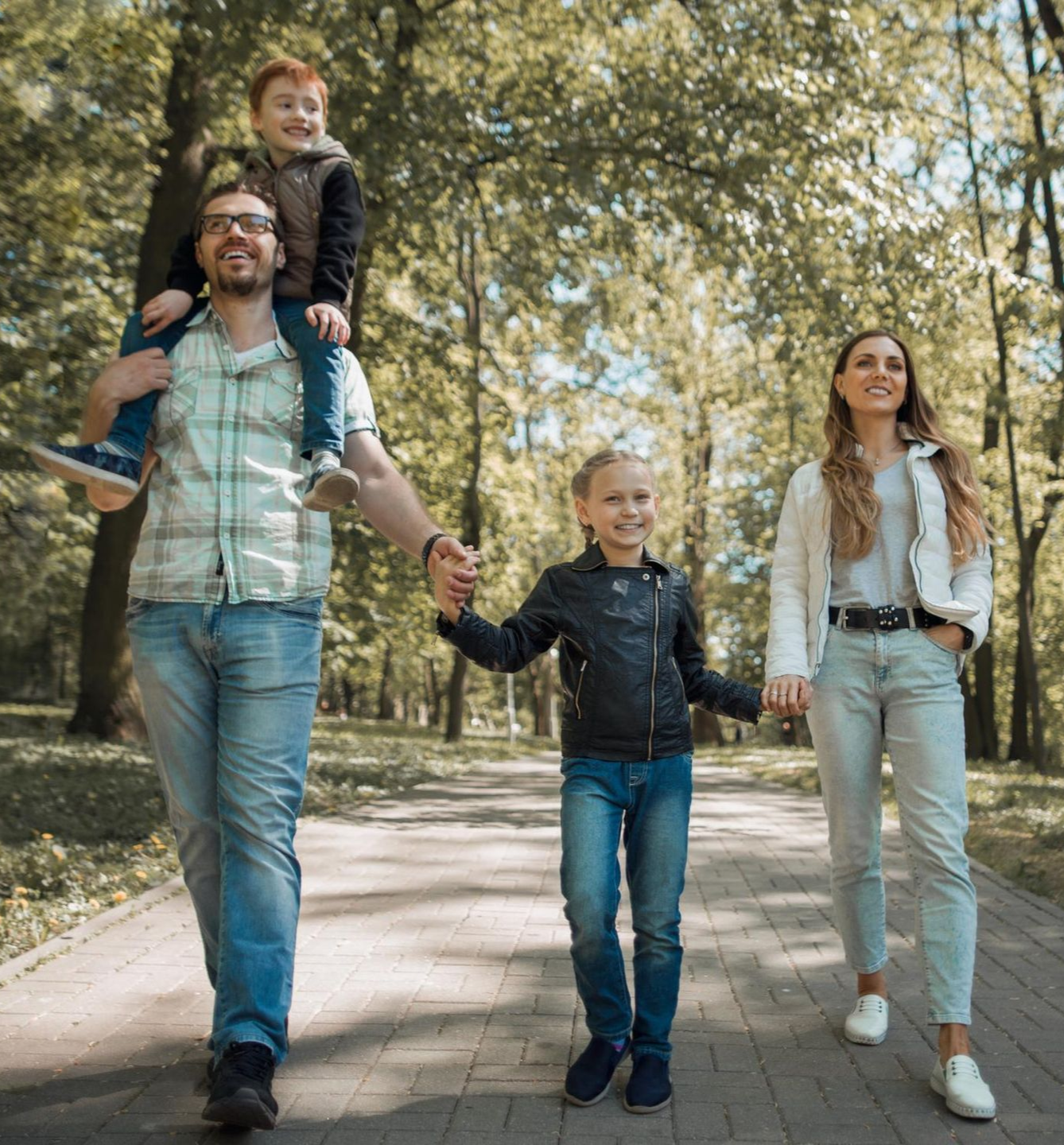 Family walking hand in hand on a tree-lined path, one child on the father's shoulders.