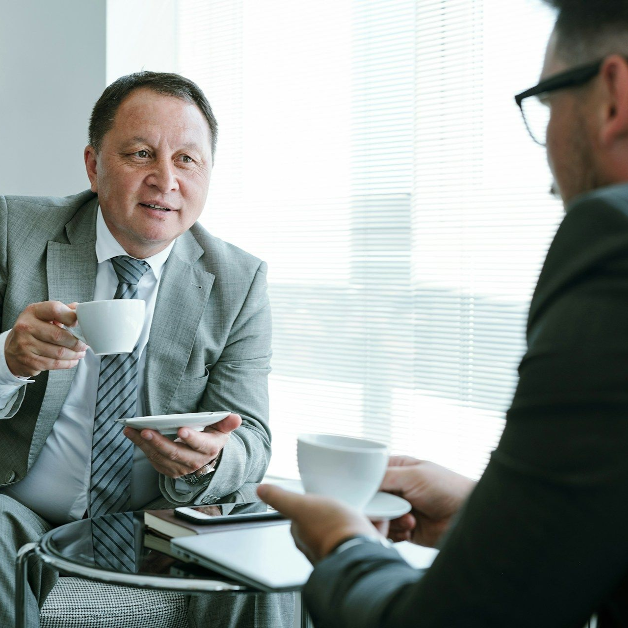 Two people in suits talking over coffee in a bright office meeting room
