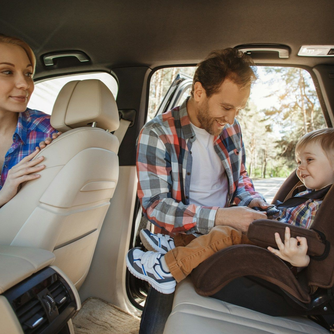 Family sitting in a car, with a man fastening a child into a car seat while a woman watches.