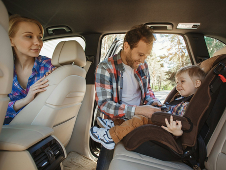 Adult buckling a child into a car seat while another passenger sits in the back of an SUV.