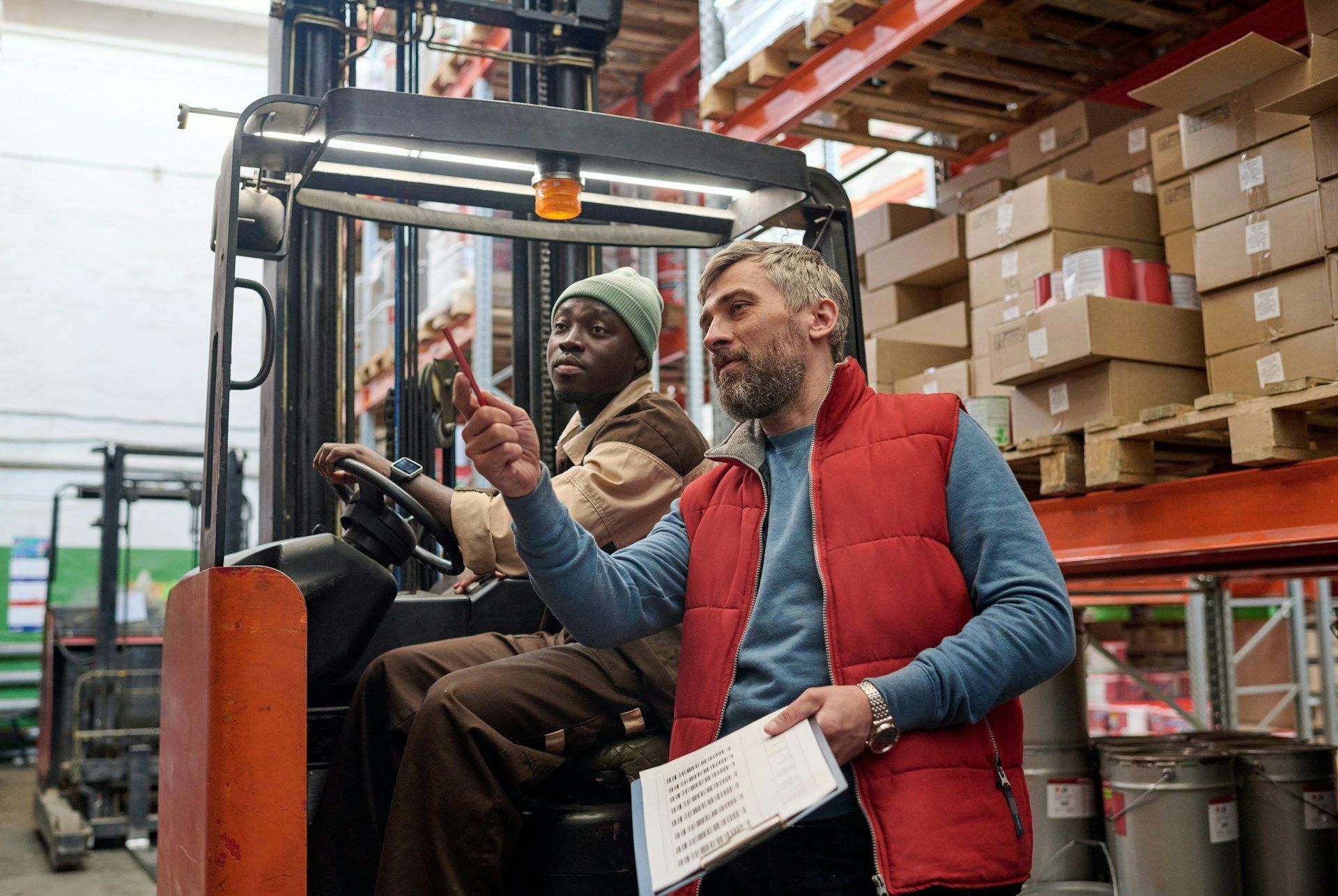 Two workers in a warehouse, one on a forklift and one holding a clipboard amid stacked boxes.