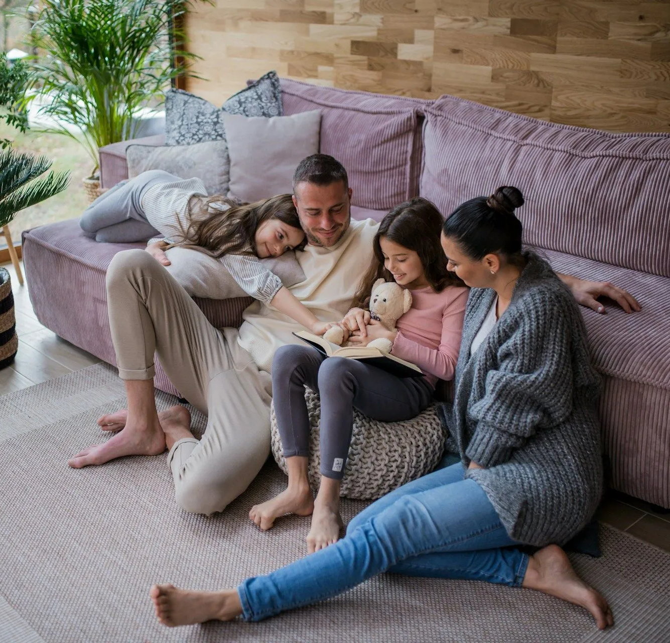 Family sitting on a couch in a cozy living room, cuddling a sleeping baby and a teddy bear.
