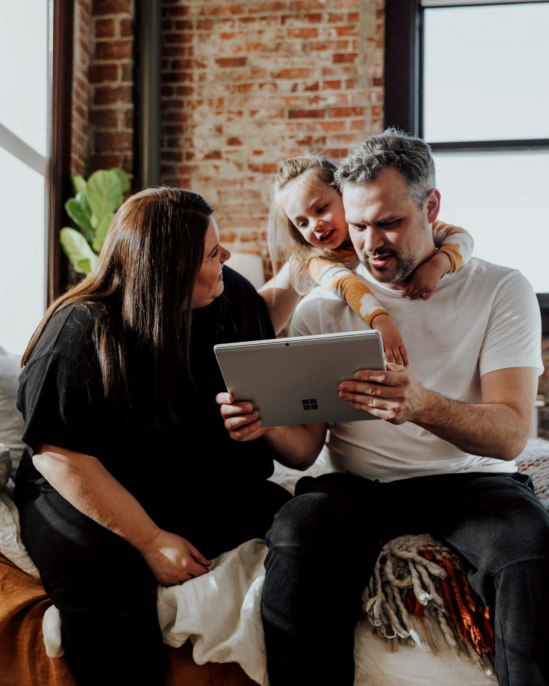 Three people sitting on a couch, looking at a tablet and smiling in a cozy brick-walled living room