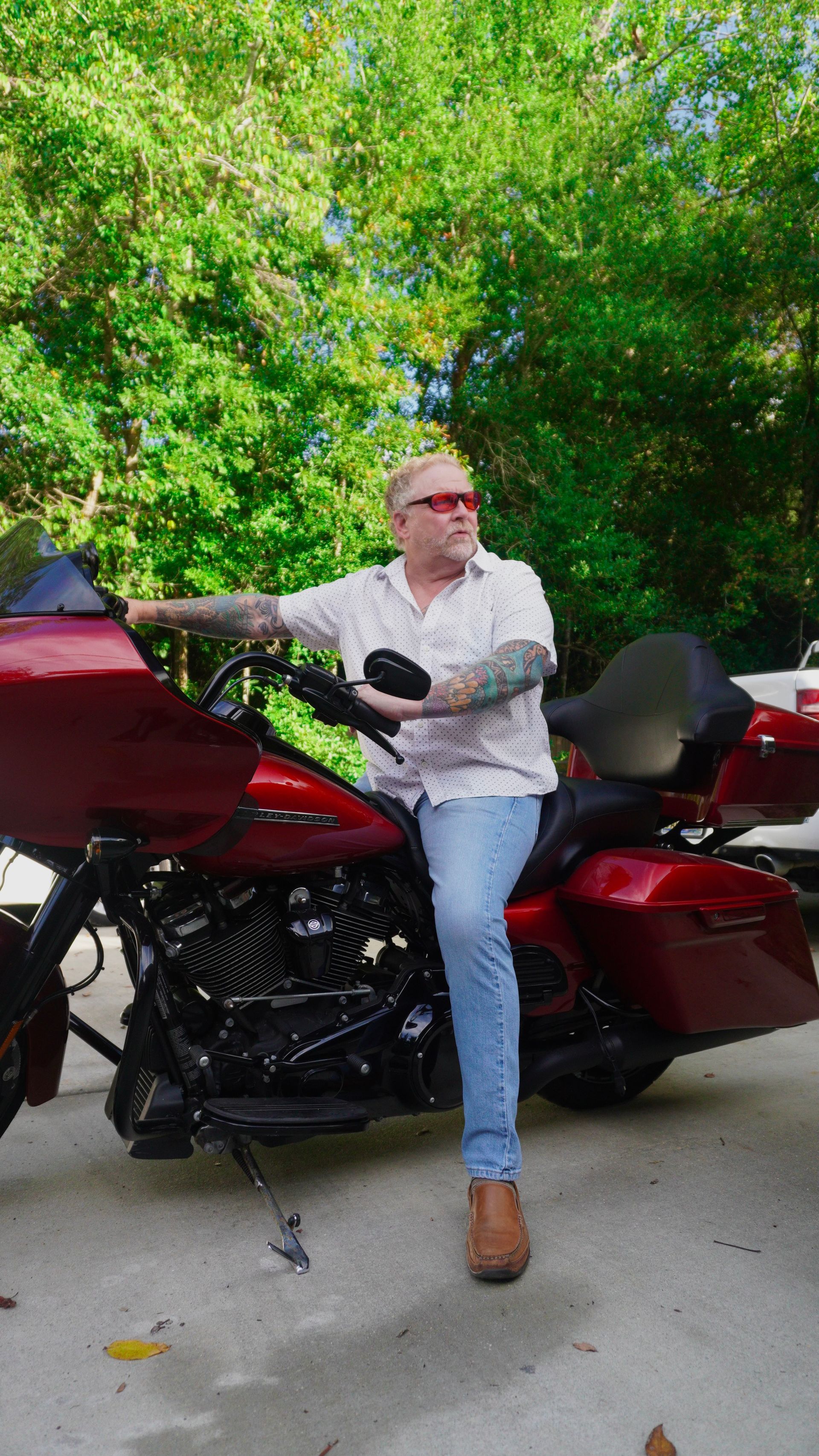 Man with sunglasses and tattoos leaning on a red motorcycle, set in a sunny, green outdoor space.