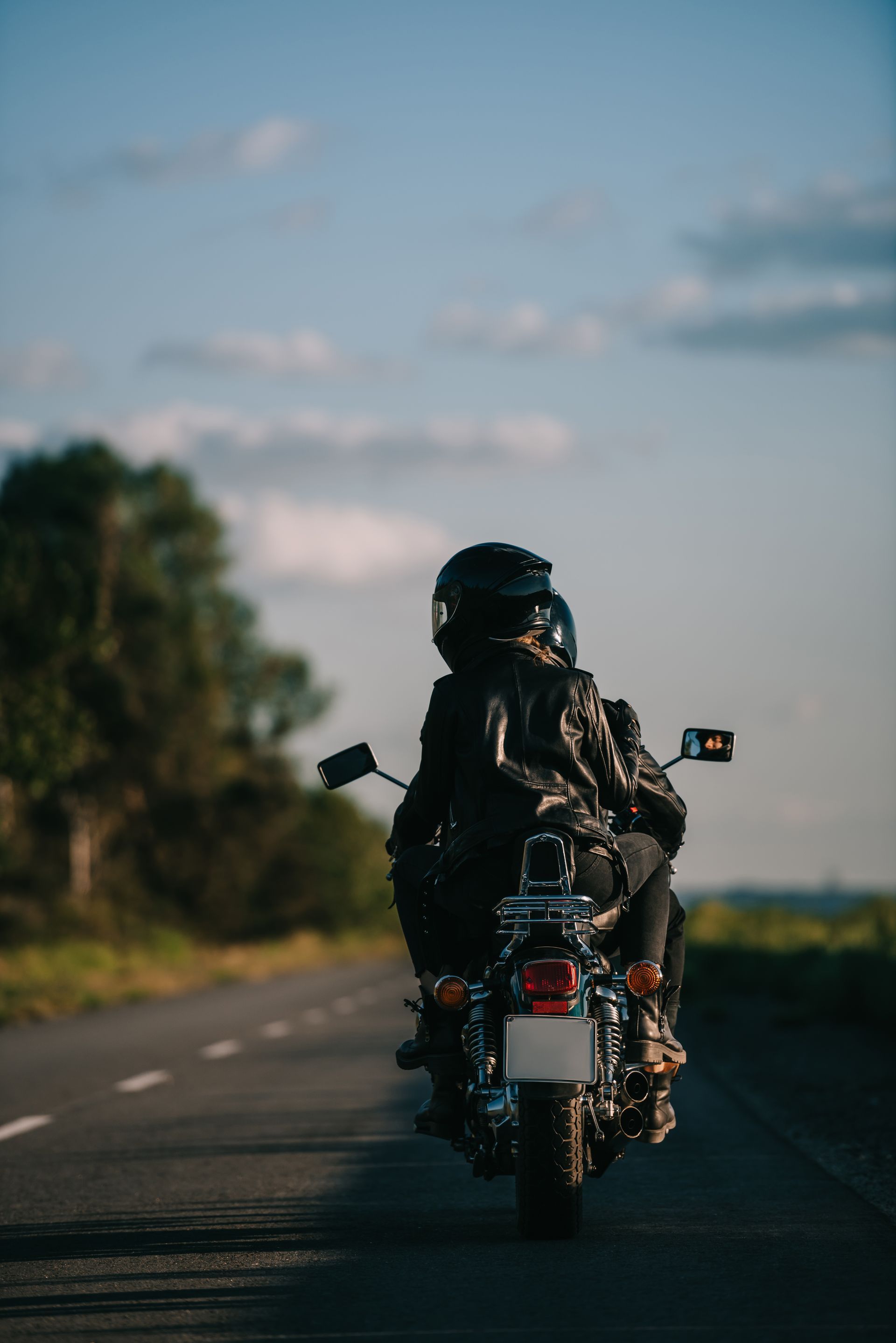 Motorcyclist on black bike riding down a road with green foliage and blue sky.