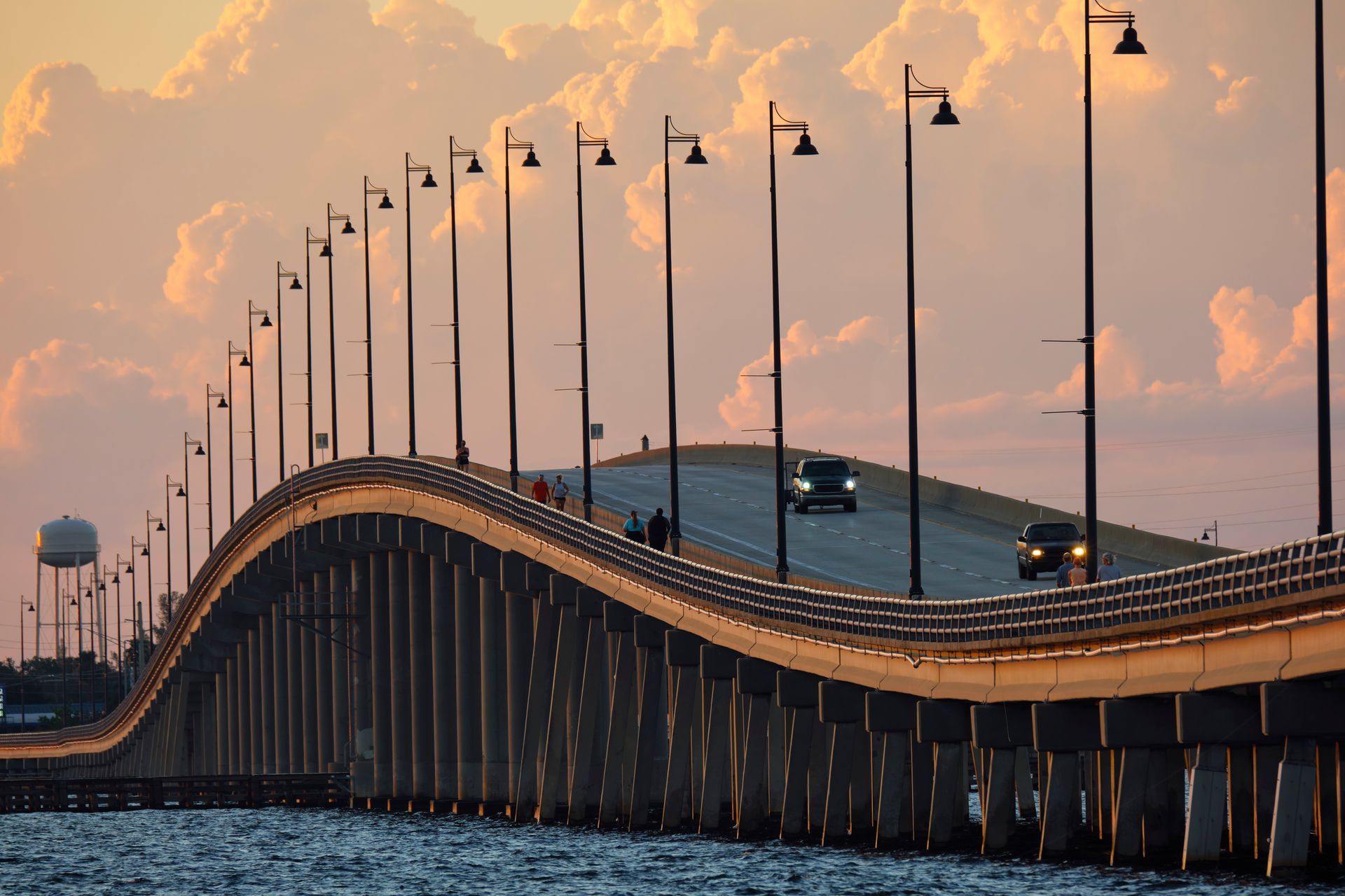 Bridge over water at sunset; cars driving, streetlights, pink and orange clouds.